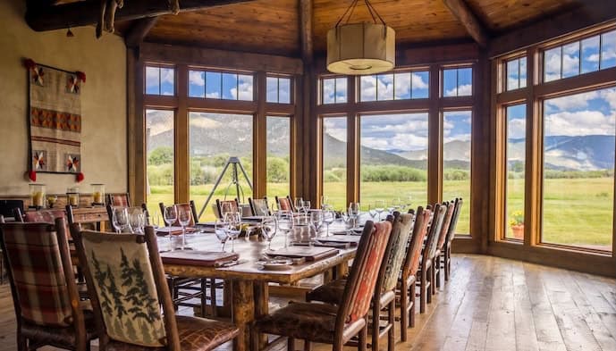 Dining room table set with breakfast spread inside a lodge with large windows overlooking open landscape and mountains.