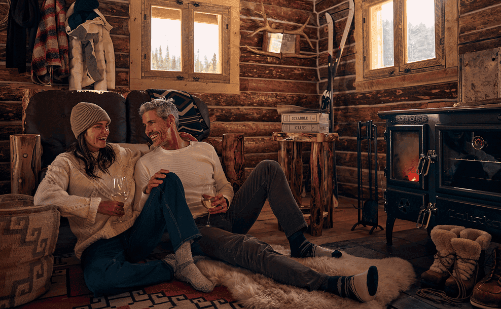 Couple relaxing by a wood stove inside a cozy log cabin with warm rustic decor.