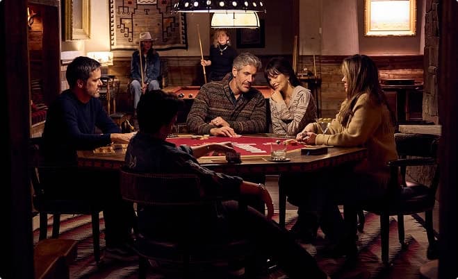Group of people playing cards around a table in a cozy lodge setting.