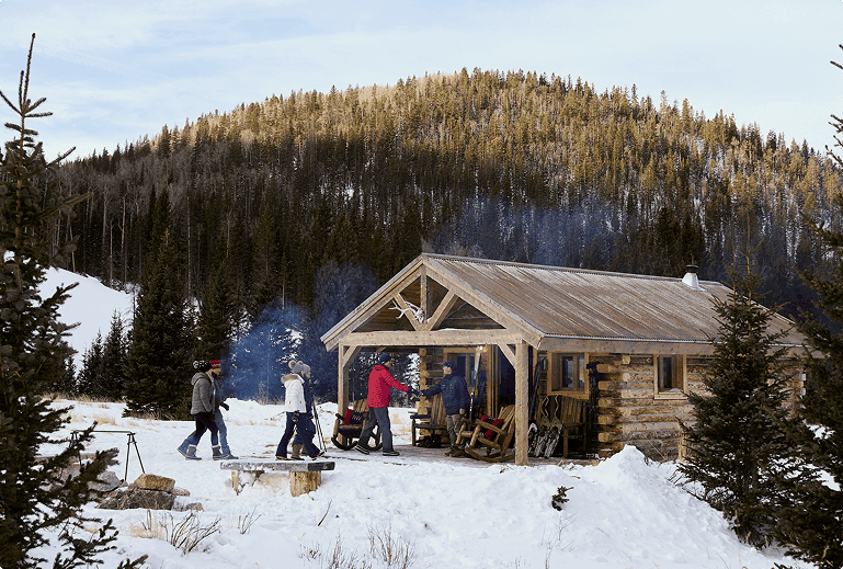 Group gathered outside a rustic cabin in a snowy forest, with mountains in the background.