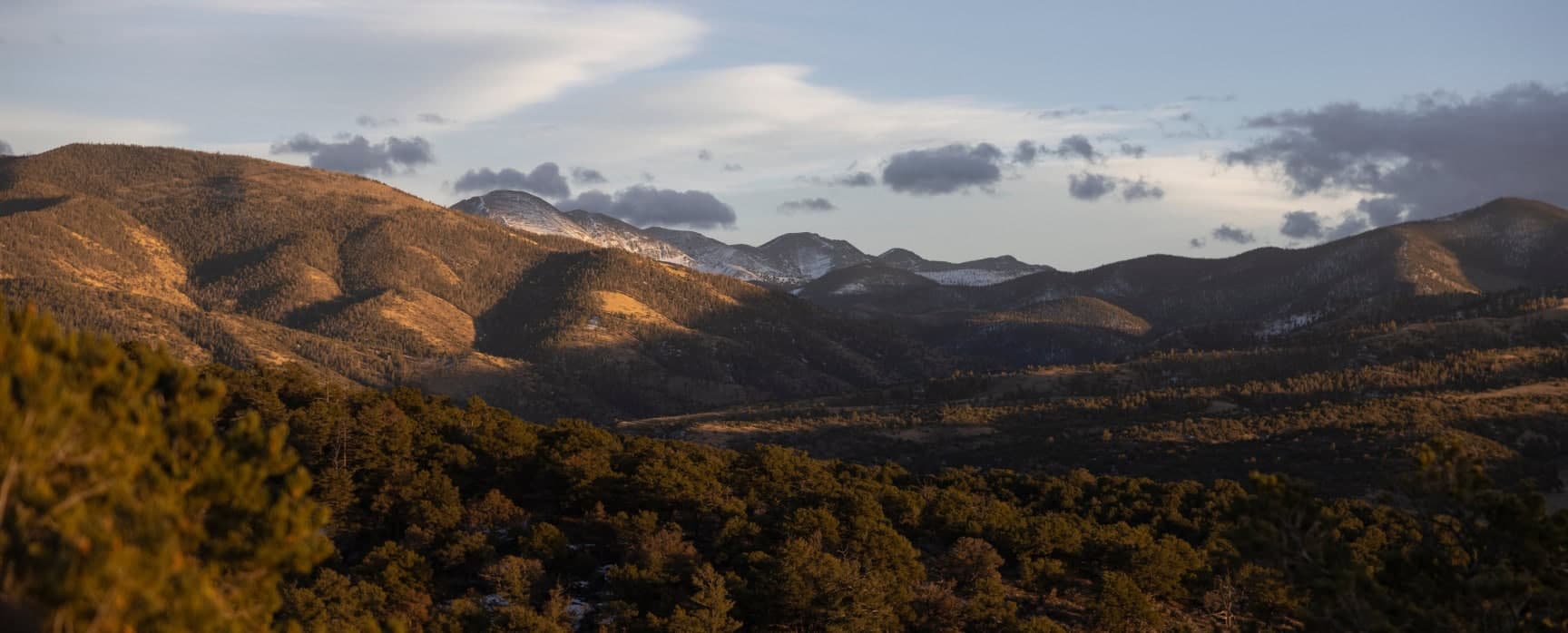 Mountain landscape at golden hour with rolling forested hills and snow-dusted peaks under soft evening light.