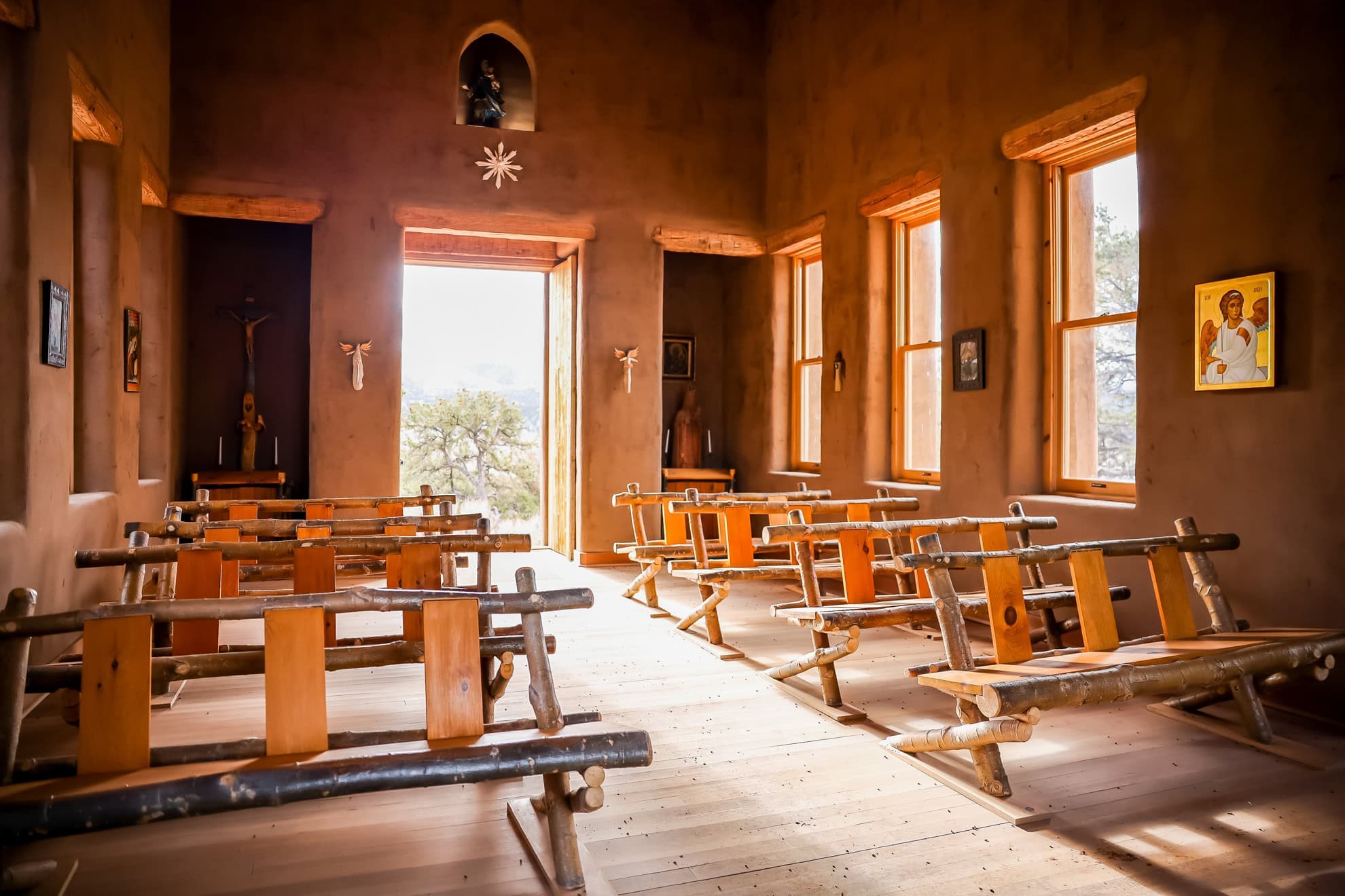 Rustic adobe chapel interior with handcrafted wooden pews, warm natural light, religious artwork, and mountain views through the open doorway at Trinchera Reserve & Lodge.
