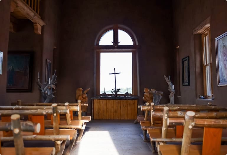 Intimate chapel ceremony space with handcrafted wooden pews, adobe walls, and a simple altar beneath an arched window at Trinchera Reserve & Lodge.