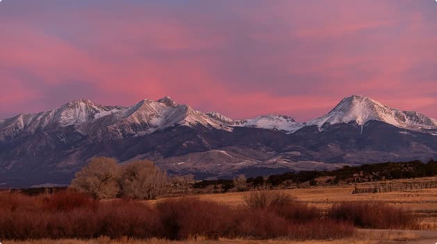 Snow-capped Sangre de Cristo Mountains glowing beneath a dramatic pink sunset sky, viewed from the expansive landscape of Trinchera Reserve & Lodge.