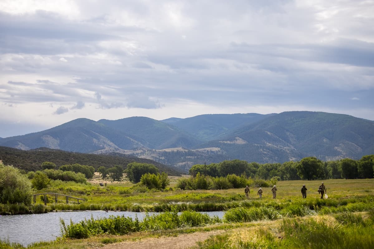 Anglers walk through a grassy meadow beside a pond, surrounded by trees and mountains under a cloudy sky.